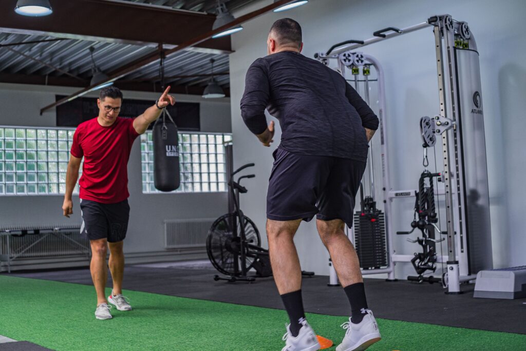 Back view of muscular man in sportswear doing exercises with trainer in modern fitness club